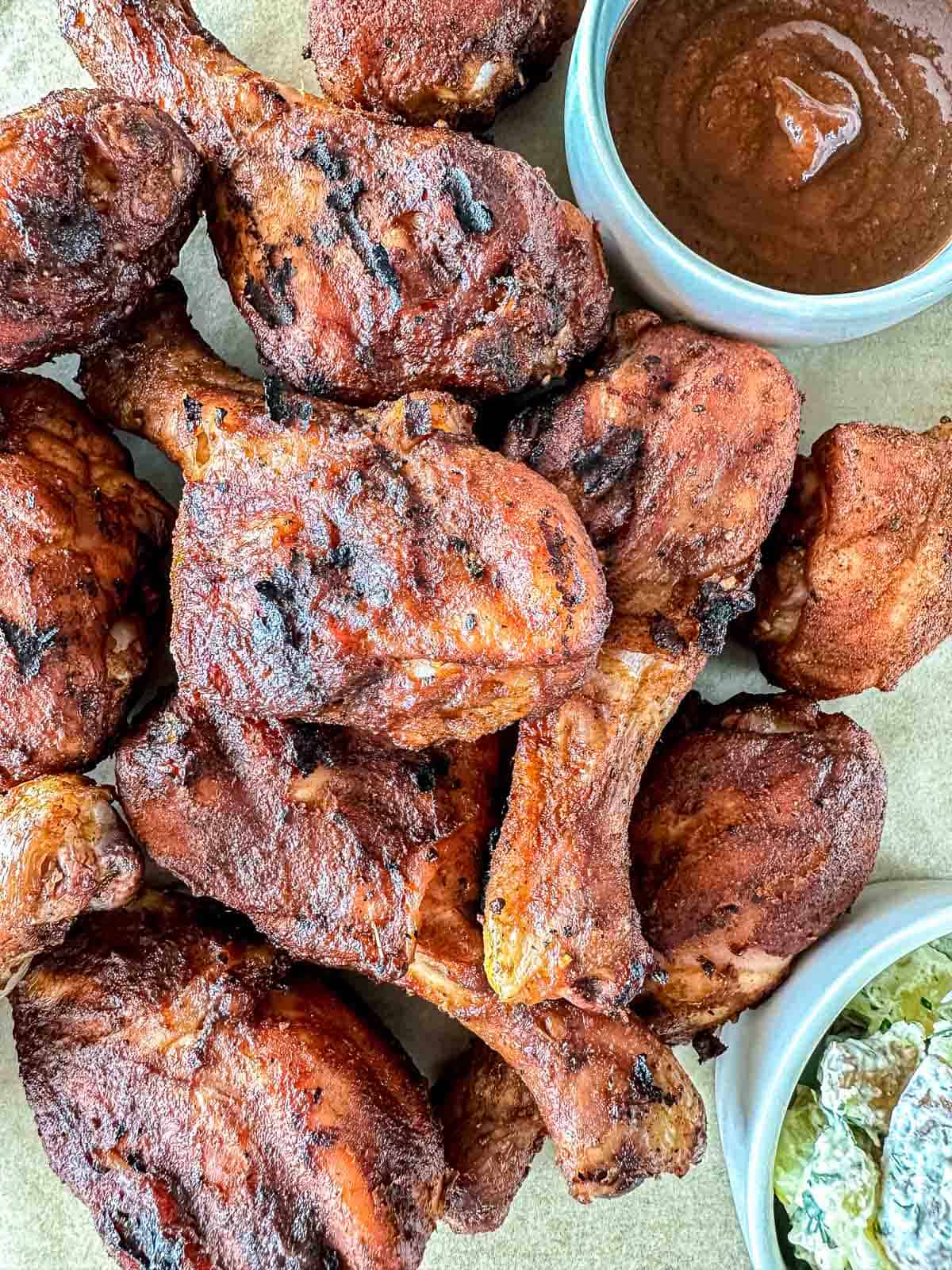 A pile of grilled barbecue chicken drumsticks on a plate, served with a bowl of barbecue sauce and a bowl of creamy coleslaw on the side.