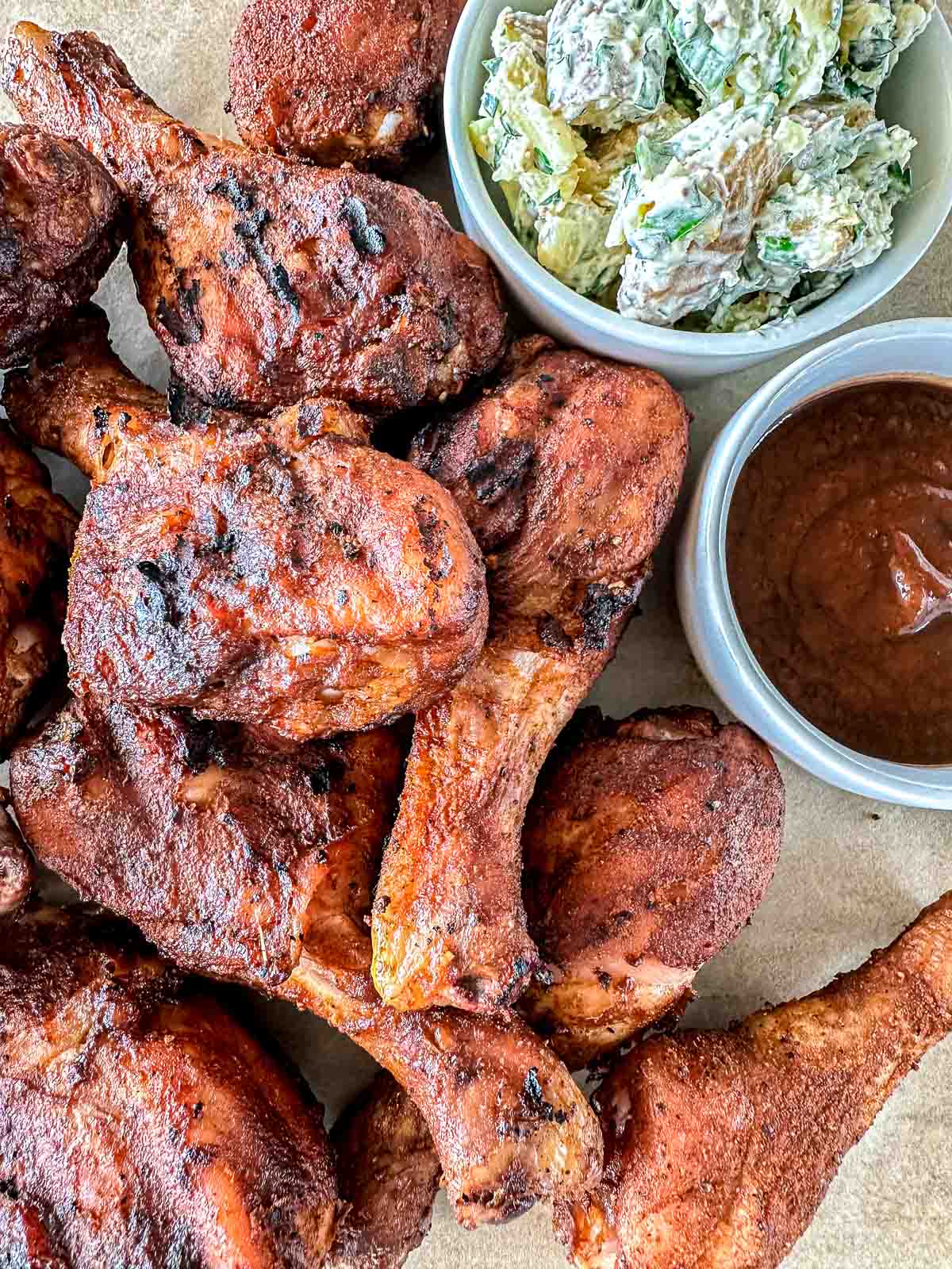 A close-up of grilled barbecue chicken drumsticks piled together, served with a bowl of creamy potato salad and a bowl of barbecue sauce on the side.