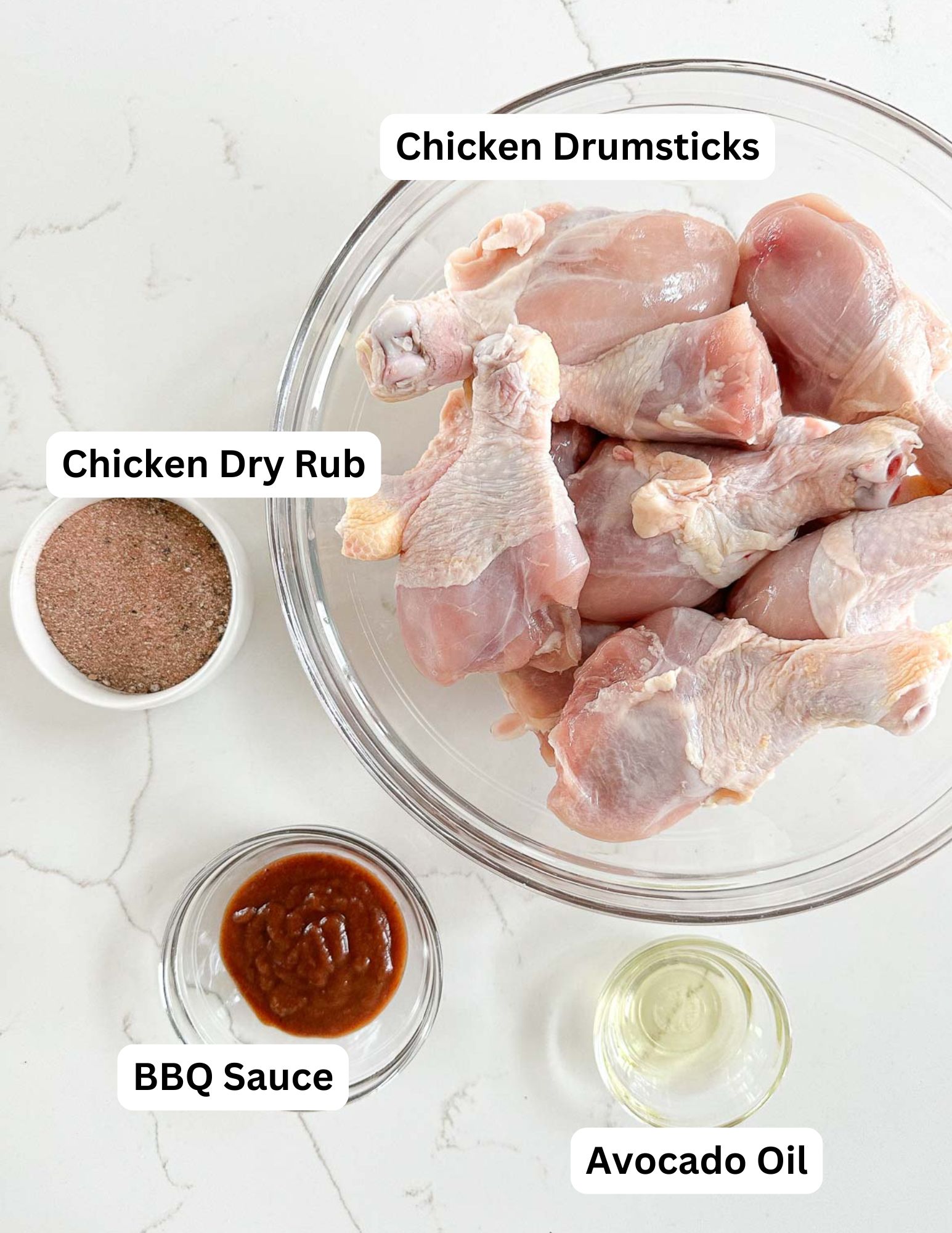A bowl of raw chicken drumsticks with labeled ingredients around it: chicken dry rub in a small bowl, BBQ sauce in a glass dish, and avocado oil in a small glass container, all on a white surface.