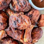 A close-up of several smoked chicken drumsticks with visible char marks, arranged on a surface next to a small cup of dark barbecue sauce.