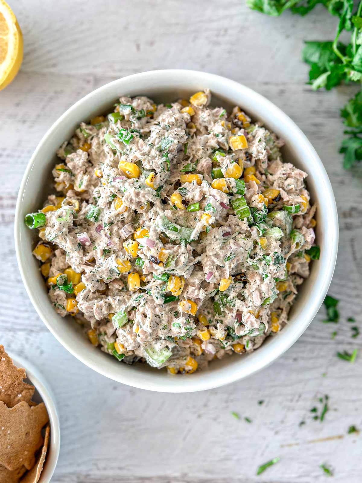 A bowl filled with tuna salad mixed with corn, chopped green onions, red onions, and herbs sits on a light wooden surface. Fresh herbs and a lemon wedge are partially visible nearby.