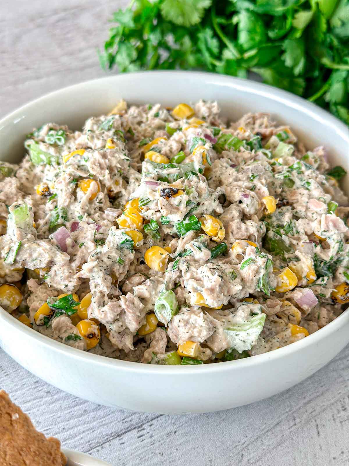 A white bowl filled with a creamy tuna salad mixed with corn, chopped green onions, red onions, cilantro, and seasonings, set on a light surface with fresh herbs in the background.