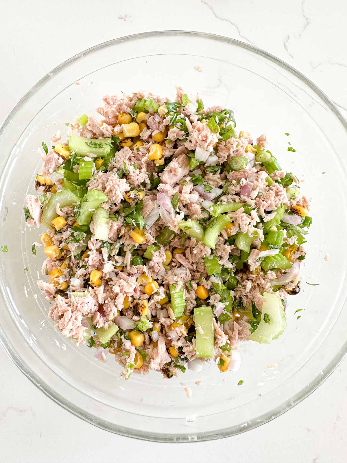 A glass bowl filled with a mixed salad containing tuna, corn, chopped celery, red onions, herbs, on a white countertop.
