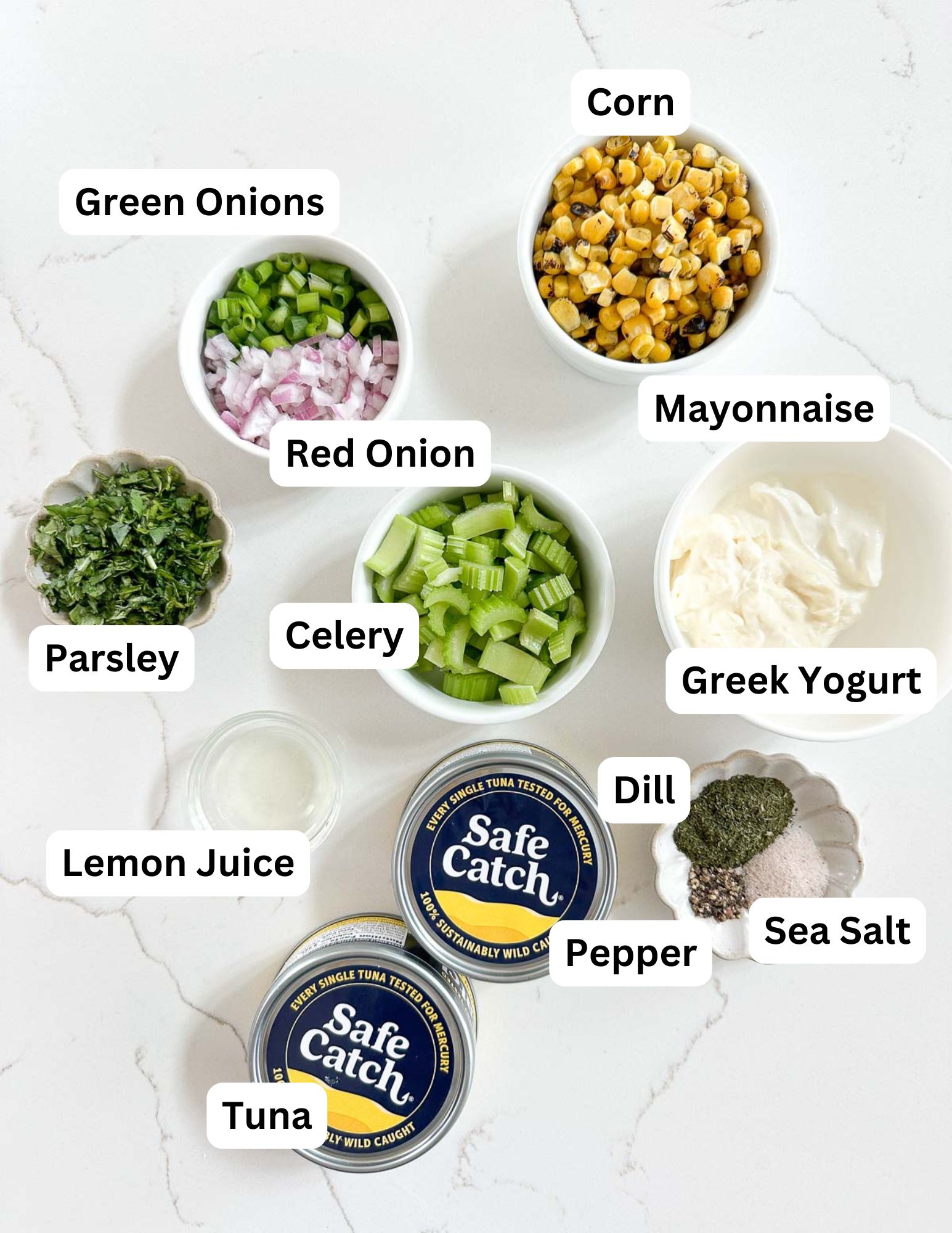 Ingredients for tuna salad are arranged on a white surface, including bowls of corn, mayonnaise, Greek yogurt, celery, red onion, green onions, parsley, lemon juice, dill, pepper, sea salt, and cans of tuna, each labeled.