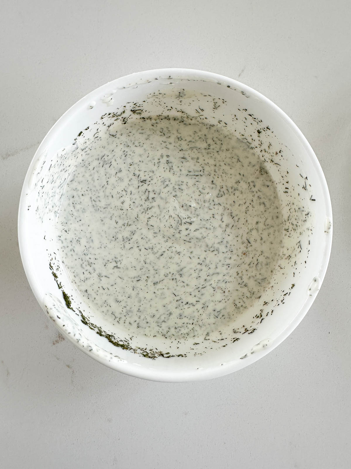A white bowl filled with creamy dressing speckled with herbs, placed on a light-colored countertop.