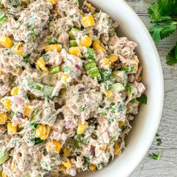A close-up of a bowl filled with tuna salad mixed with corn, chopped red onion, celery, and fresh herbs, on a light wooden surface with parsley leaves nearby.