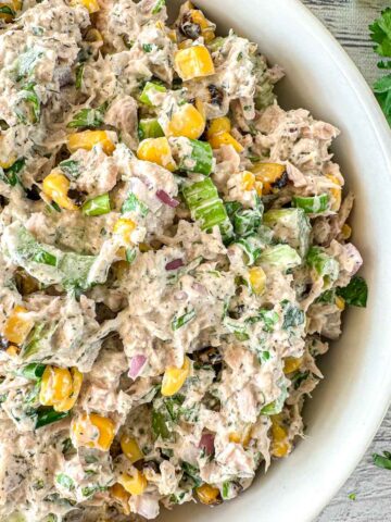 A close-up of a bowl filled with tuna salad mixed with corn, chopped red onion, celery, and fresh herbs, on a light wooden surface with parsley leaves nearby.