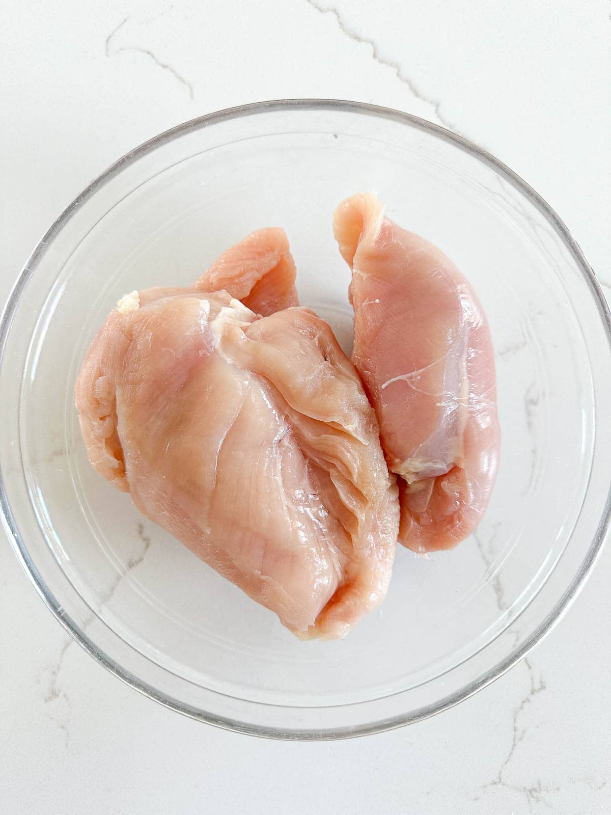 Raw chicken breasts in a clear glass bowl, placed on a white marble countertop.