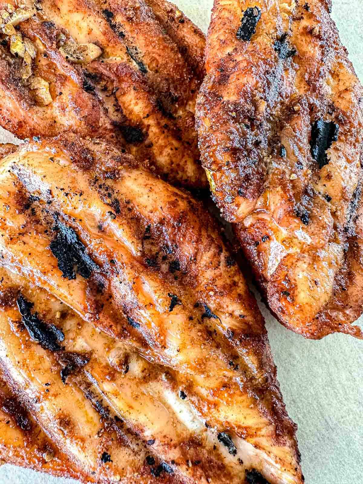 Close-up of three grilled chicken breasts with visible grill marks and a golden-brown, slightly charred exterior, placed on a white surface.