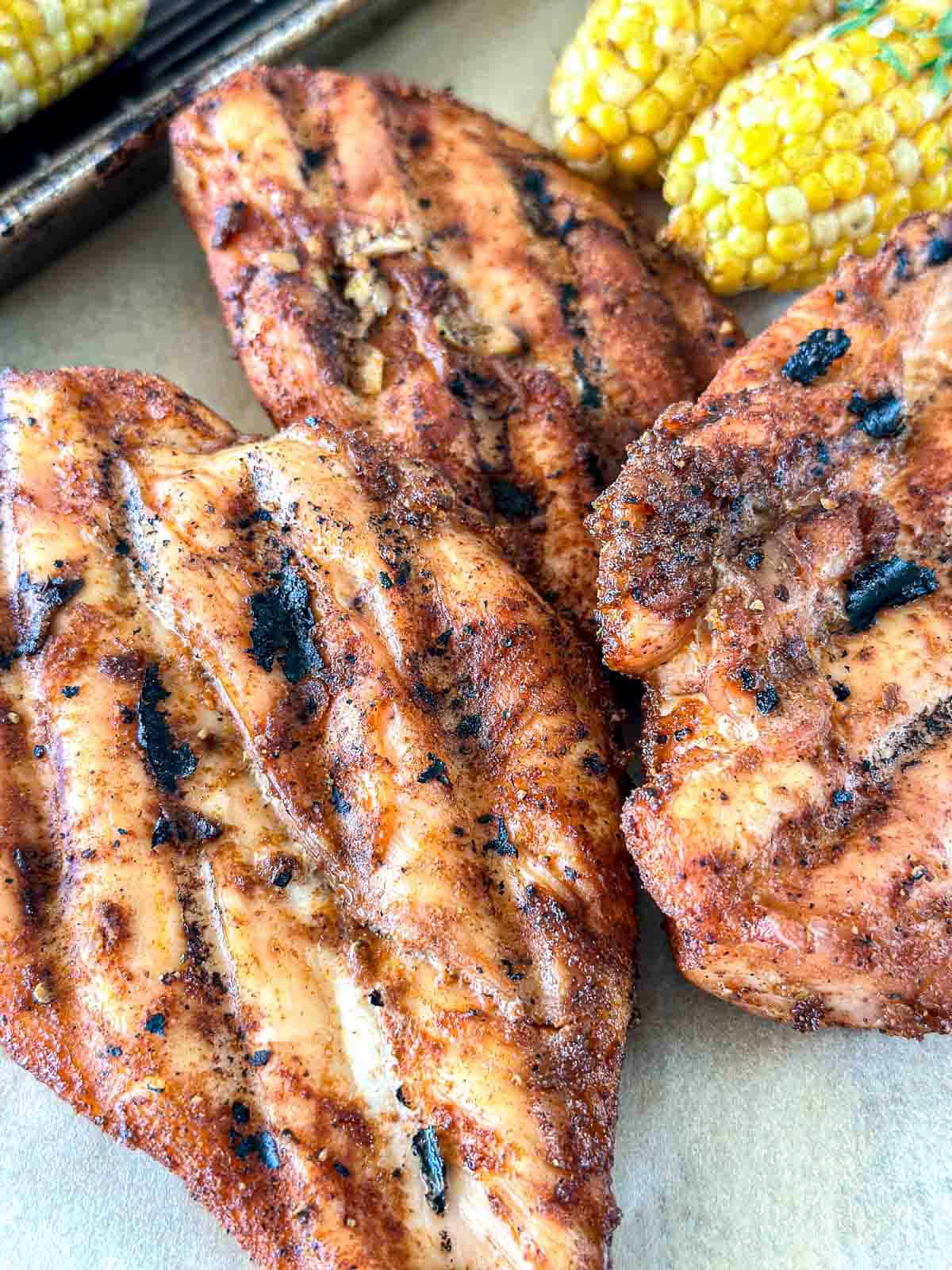 Traeger smoked chicken breasts with visible grill marks are served on a tray next to pieces of corn on the cob. The food appears juicy and freshly cooked.