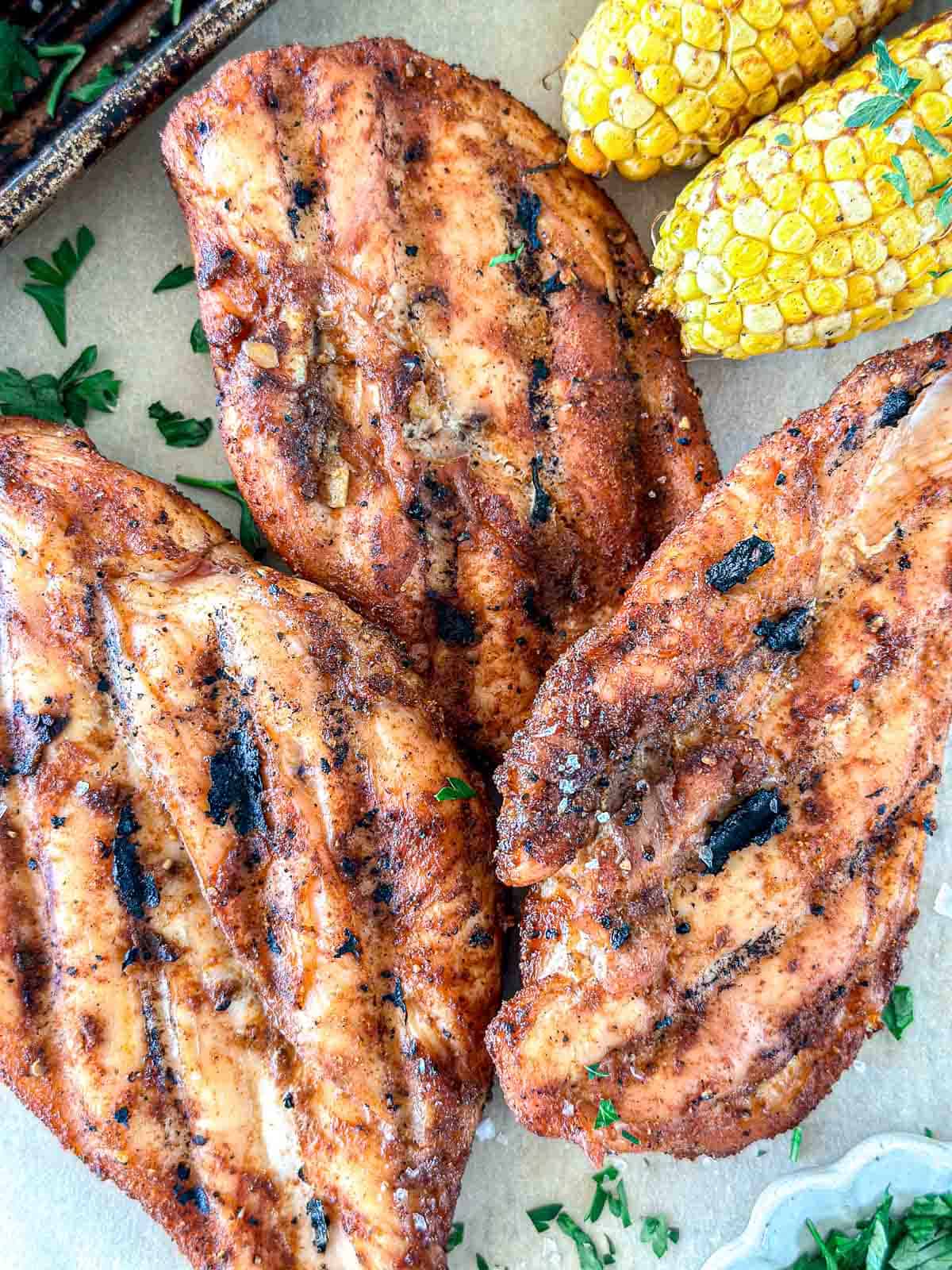 Three grilled chicken breasts with visible grill marks are arranged on a surface next to two pieces of corn on the cob and sprinkled chopped herbs. The food looks freshly cooked and seasoned.
