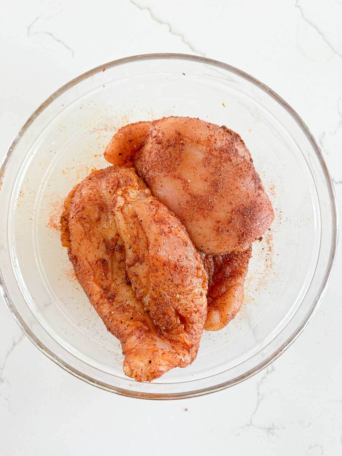 Raw chicken breasts coated with spices and seasonings in a clear glass bowl, placed on a white countertop.