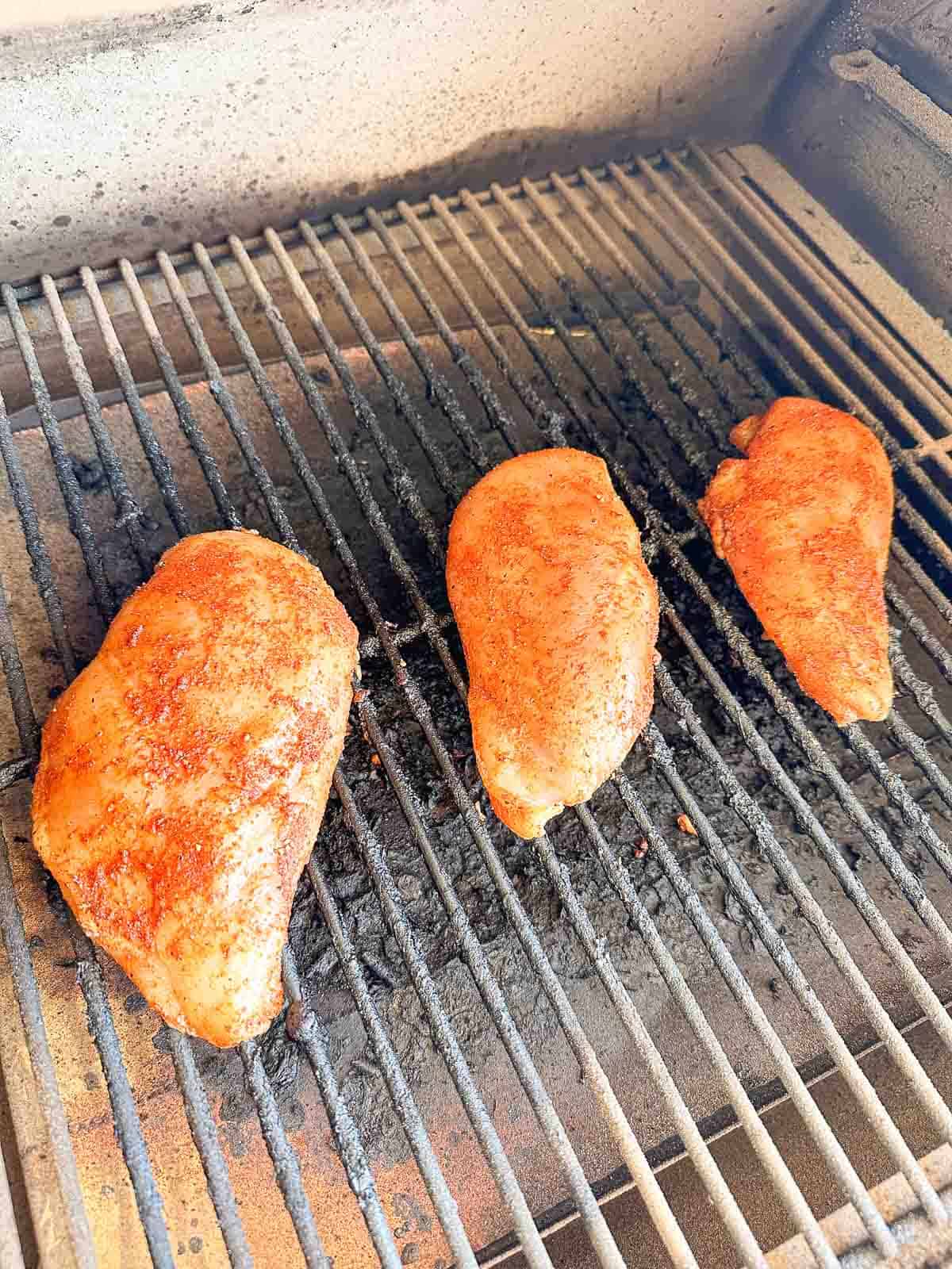 Three seasoned chicken breasts cooking on a grill with visible grill marks and char lines, set against a light-colored concrete background.