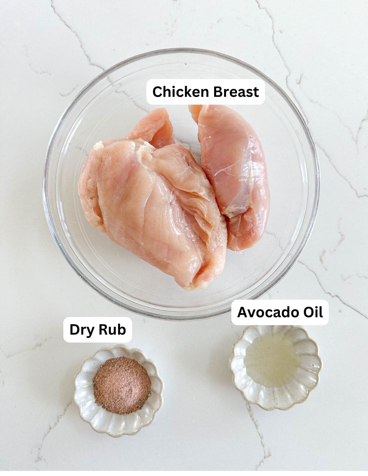 Overhead view of two raw chicken breasts in a glass bowl, with a small dish of dry rub seasoning and a small dish of avocado oil placed below on a white surface. Labels identify each item.