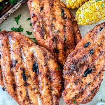 Three smoked chicken breasts with char marks are arranged on a surface, seasoned with spices. In the background, two pieces of grilled corn on the cob and some fresh herbs are visible.