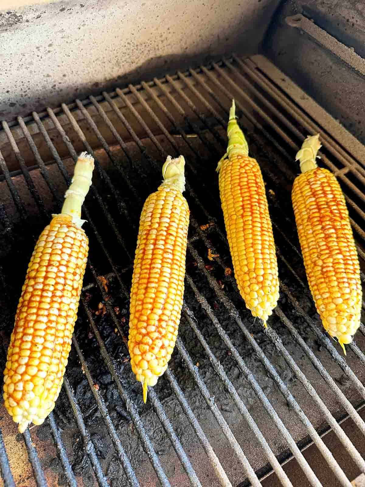 Four ears of corn with husks partially removed are being grilled, lined up on a barbecue grill with visible char marks on the kernels.