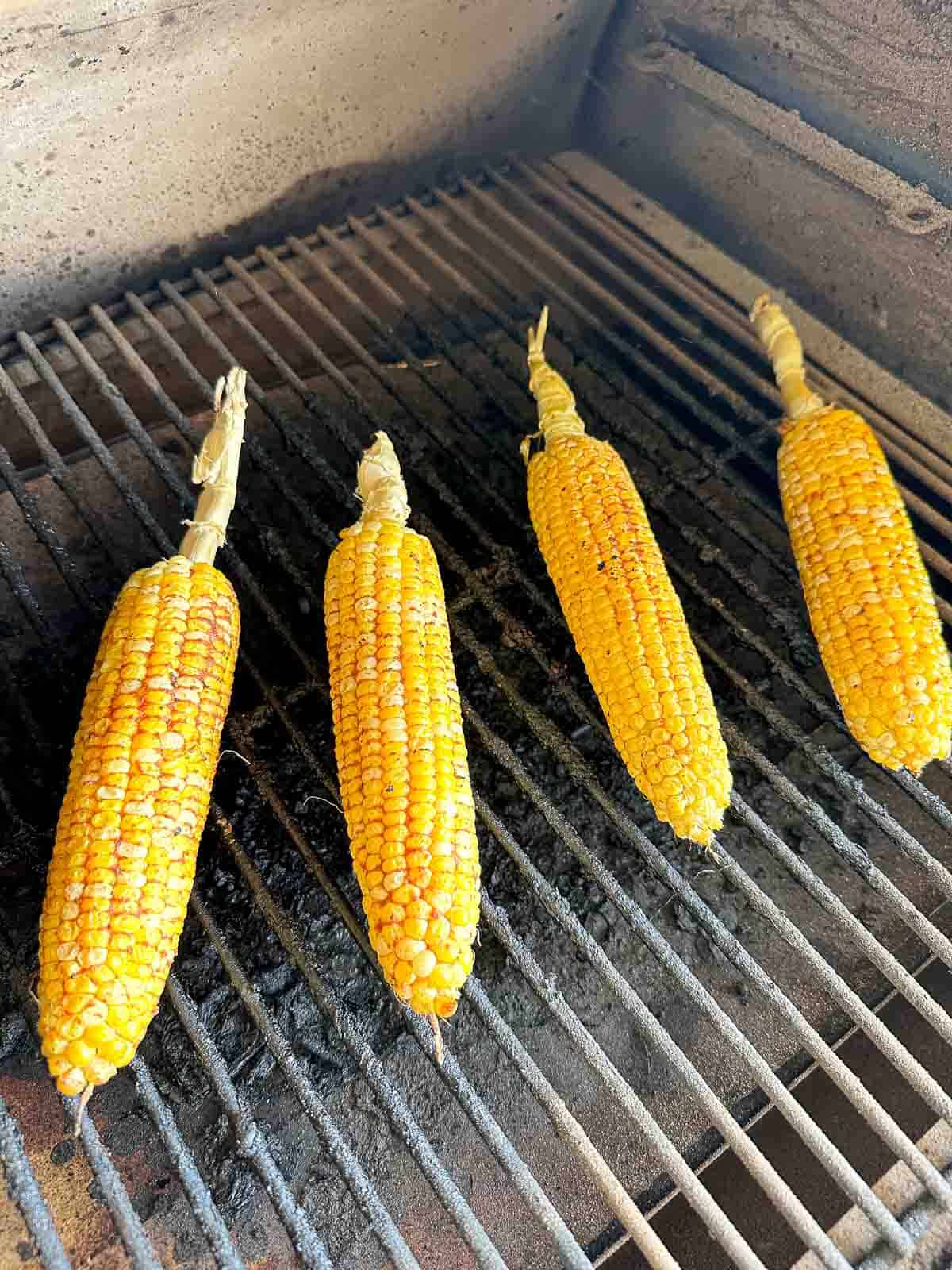 Four ears of corn, still in husks at the top, are grilling on a barbecue grill. The corn kernels are yellow with some char marks from the grill. The grill grates are visible beneath them.