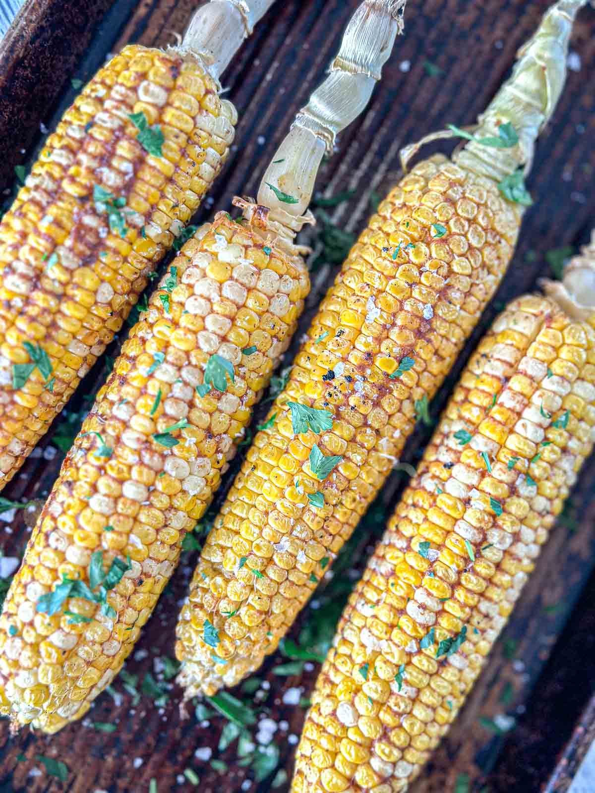 Four smoked corn cobs sprinkled with chopped herbs and coarse salt, arranged side by side on a wooden surface.