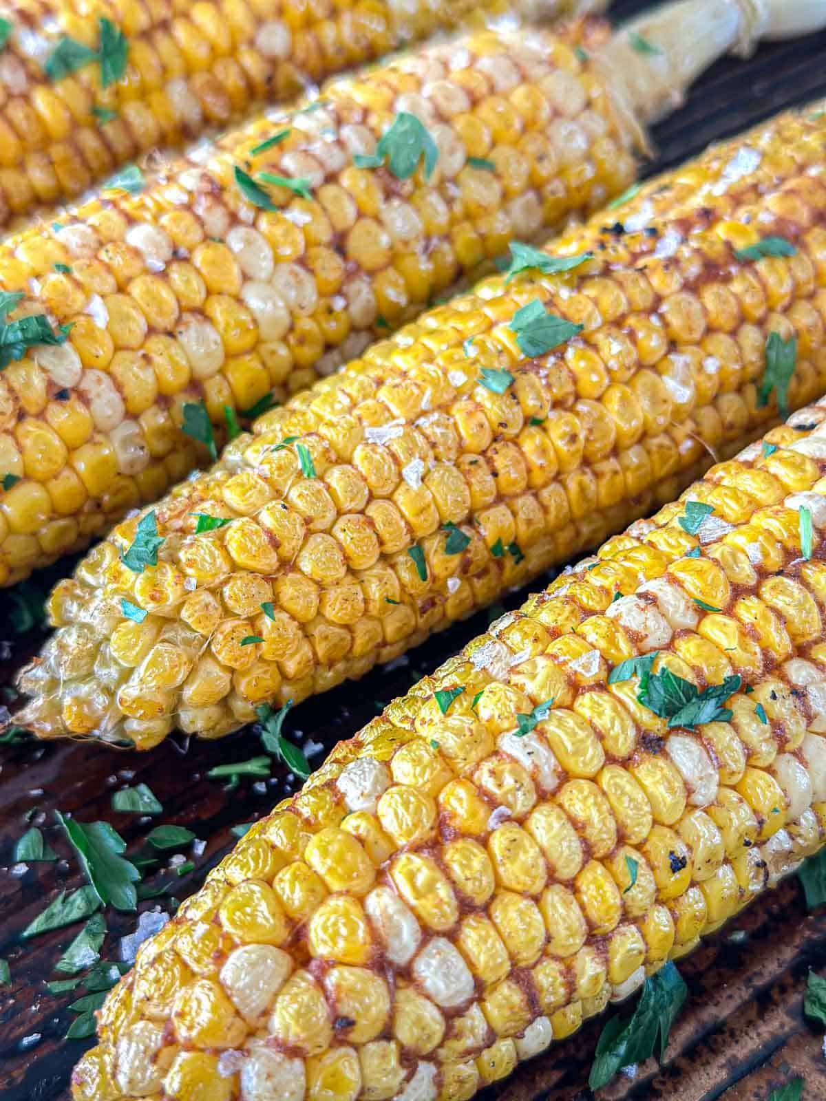 Smoked corn on the cob with char marks, topped with chopped fresh herbs and a sprinkle of salt, arranged closely together on a wooden surface.