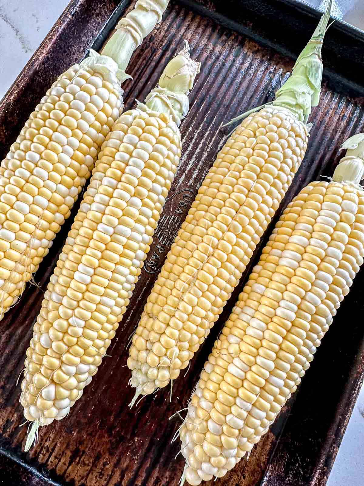Four ears of fresh corn with husks partially removed are arranged side by side on a dark, ridged baking sheet.