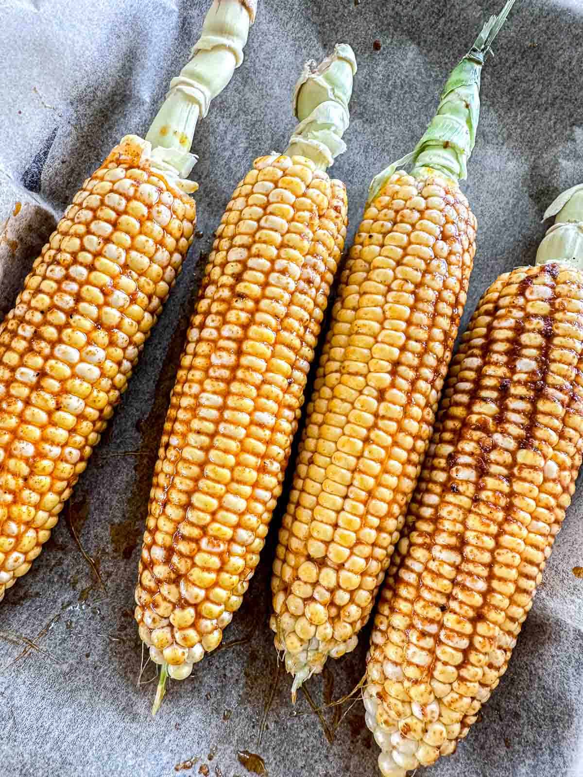 Four ears of corn with husks partially removed, arranged on a parchment-lined surface, sprinkled with a reddish-brown seasoning.