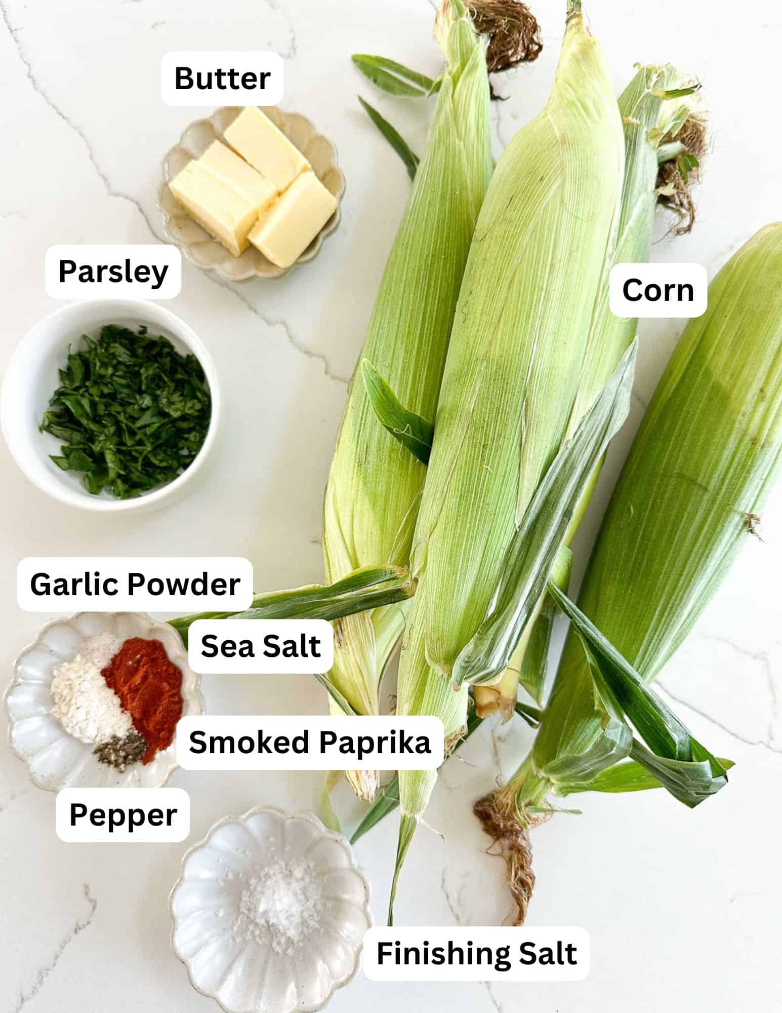 Three ears of corn in husks with small bowls of labeled ingredients: butter, parsley, garlic powder, sea salt, smoked paprika, pepper, and finishing salt, arranged on a white surface.