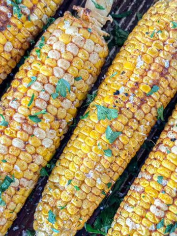 Smoked corn on the cob topped with fresh chopped herbs and a sprinkle of coarse salt, arranged closely together on a dark surface.