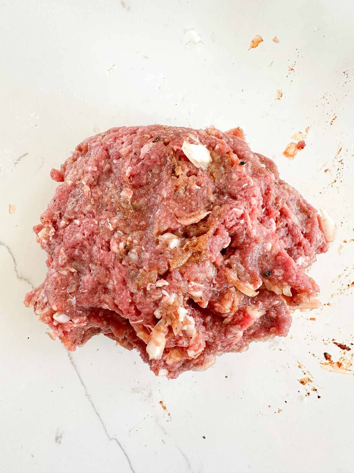 A mound of raw ground meat mixed with seasonings sits on a white countertop, with some scattered bits and spices around it.