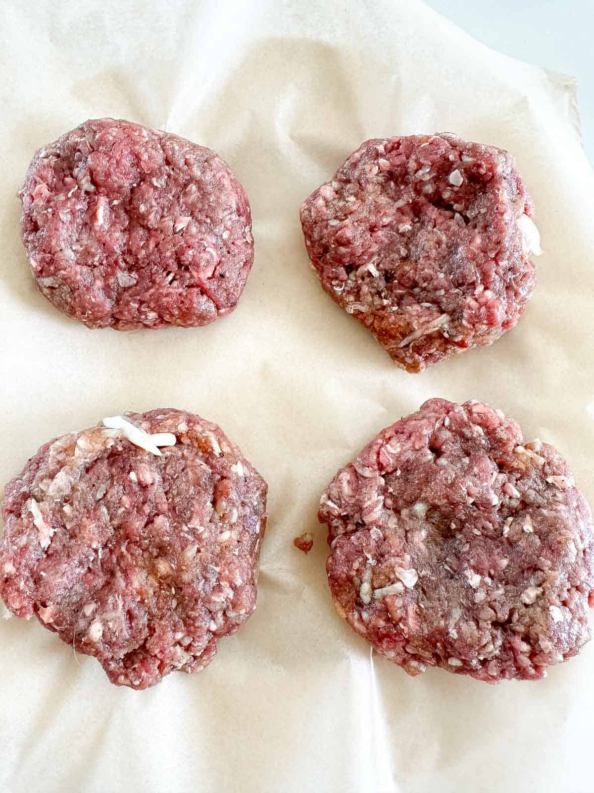 Four raw hamburger patties with visible pieces of onion are arranged on a sheet of parchment paper, ready to be cooked.