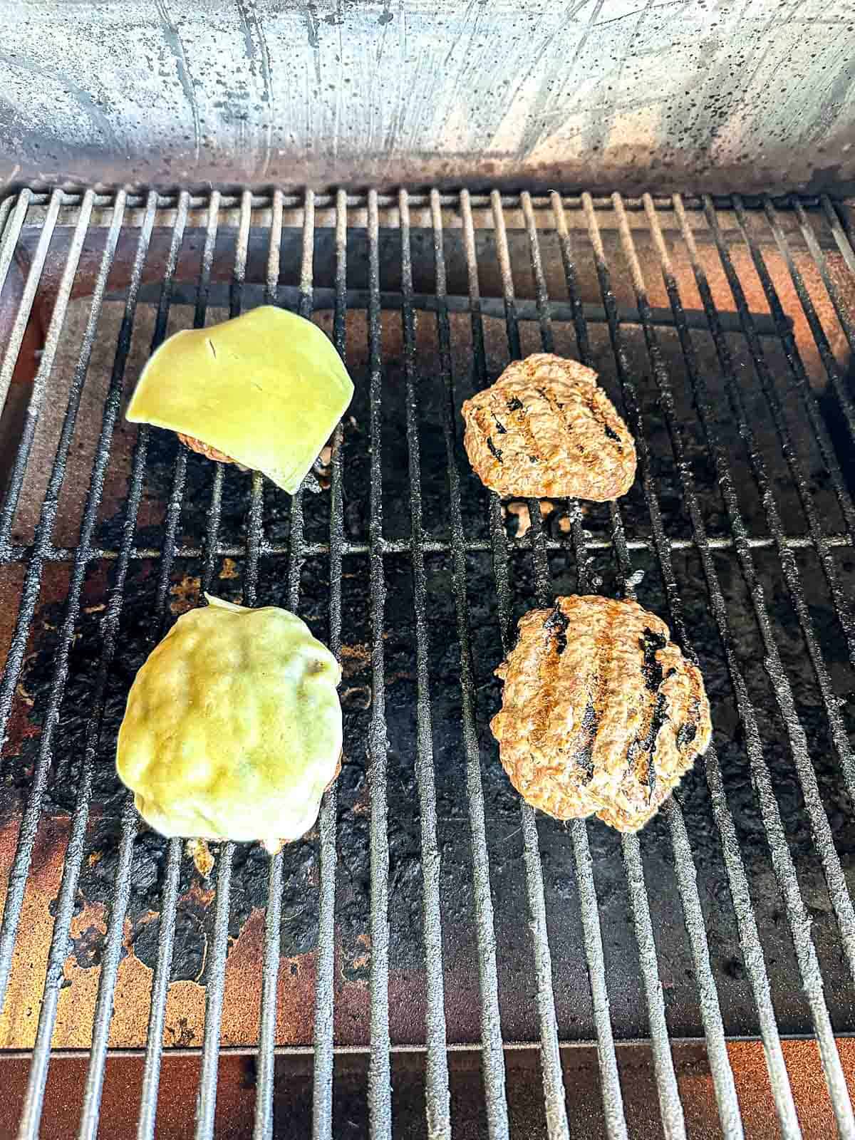 Four grilled burger patties are cooking on a grill; two are topped with melted cheese, while the other two are plain with visible grill marks. The grill surface appears slightly charred.
