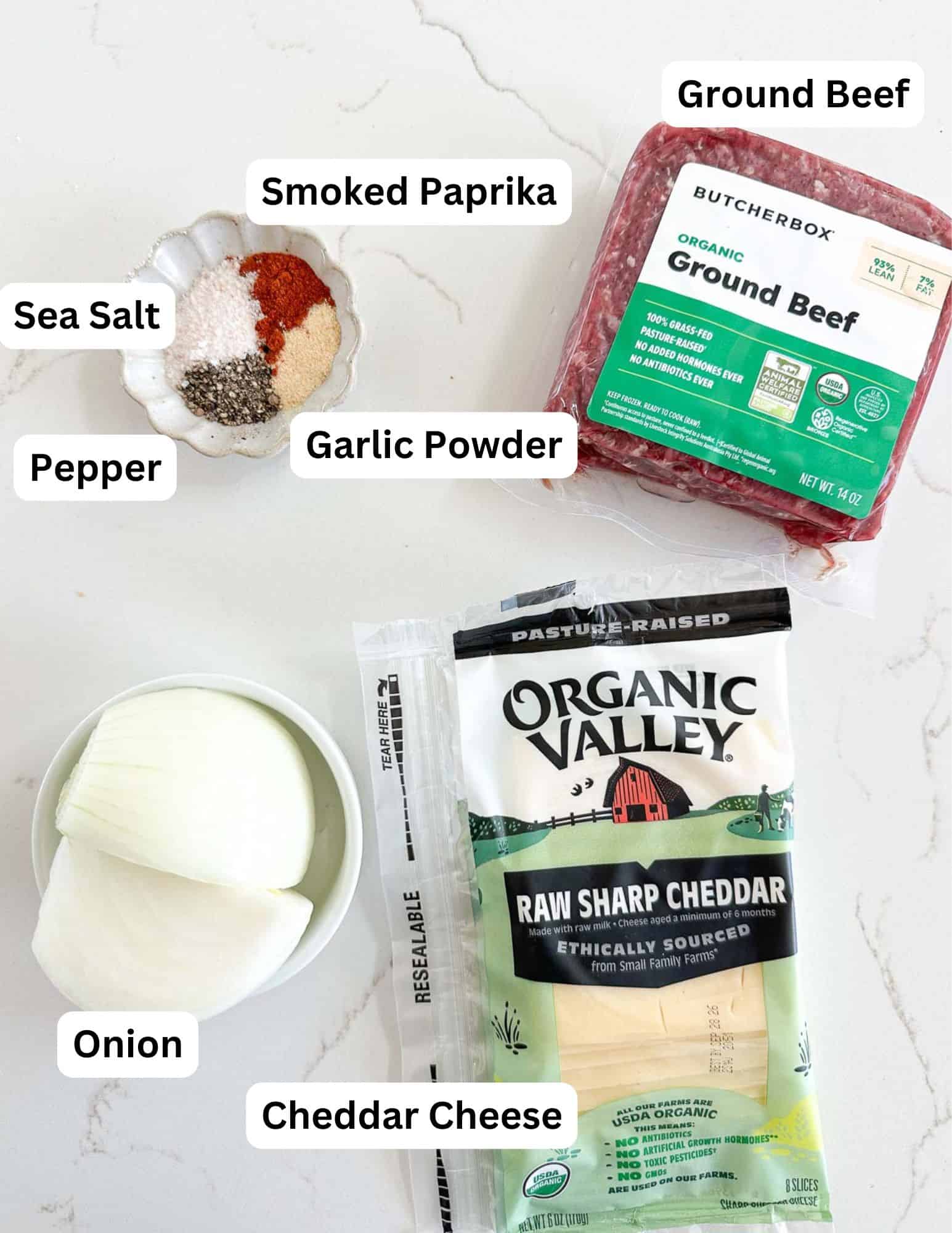 Labeled ingredients on a white surface: sea salt, pepper, smoked paprika, garlic powder in a dish, a package of ground beef, a wedge of onion, and a package of raw sharp cheddar cheese.