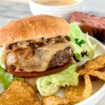 A grilled burger with lettuce, tomato, caramelized onions, and melted cheese in a bun, served on a plate with potato chips. A bowl of sauce and a slice of tomato are in the background.