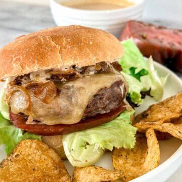 A grilled burger with lettuce, tomato, caramelized onions, and melted cheese in a bun, served on a plate with potato chips. A bowl of sauce and a slice of tomato are in the background.