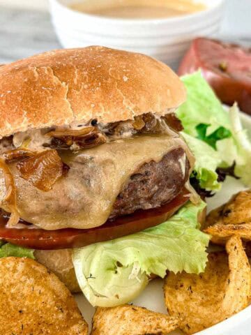 A grilled burger with lettuce, tomato, caramelized onions, and melted cheese in a bun, served on a plate with potato chips. A bowl of sauce and a slice of tomato are in the background.