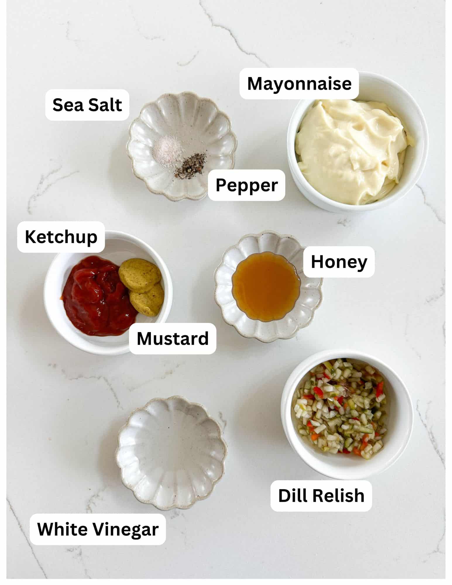 Seven small bowls with labeled ingredients for a sauce: sea salt, pepper, mayonnaise, ketchup, mustard, honey, dill relish, and white vinegar, arranged on a white surface.