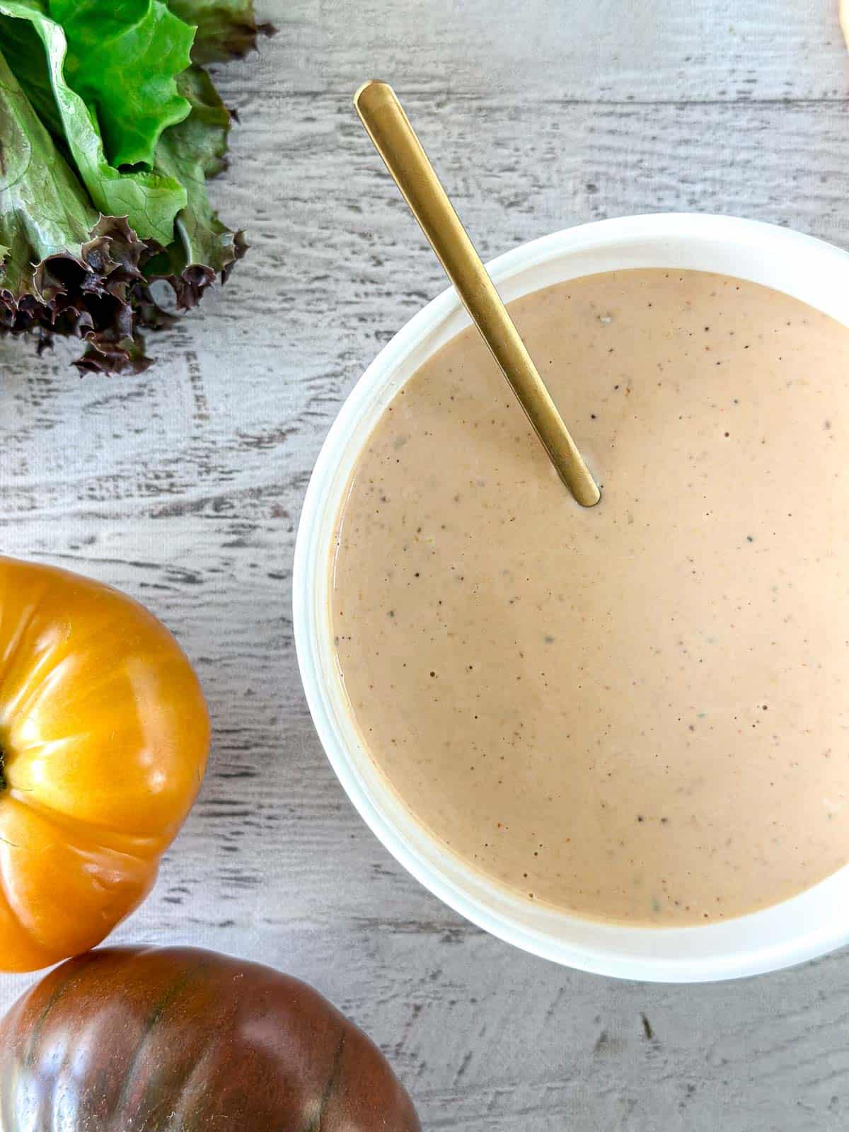 A bowl of In-N-Out sauce with a spoon in it, surrounded by leafy greens and two heirloom tomatoes on a light wooden surface.