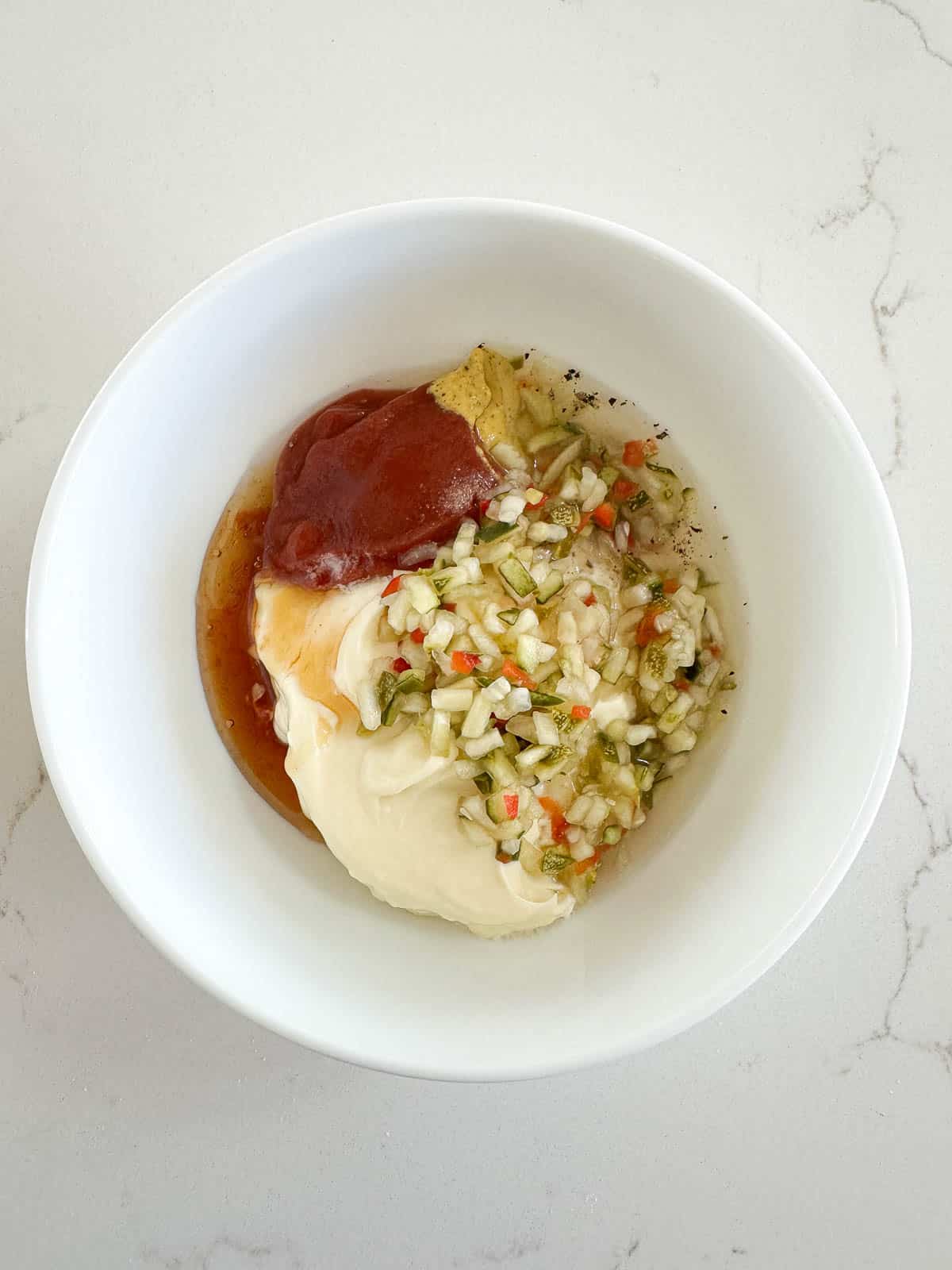 A white bowl containing mayonnaise, ketchup, chopped pickles, onions, and seasonings, placed on a white countertop.