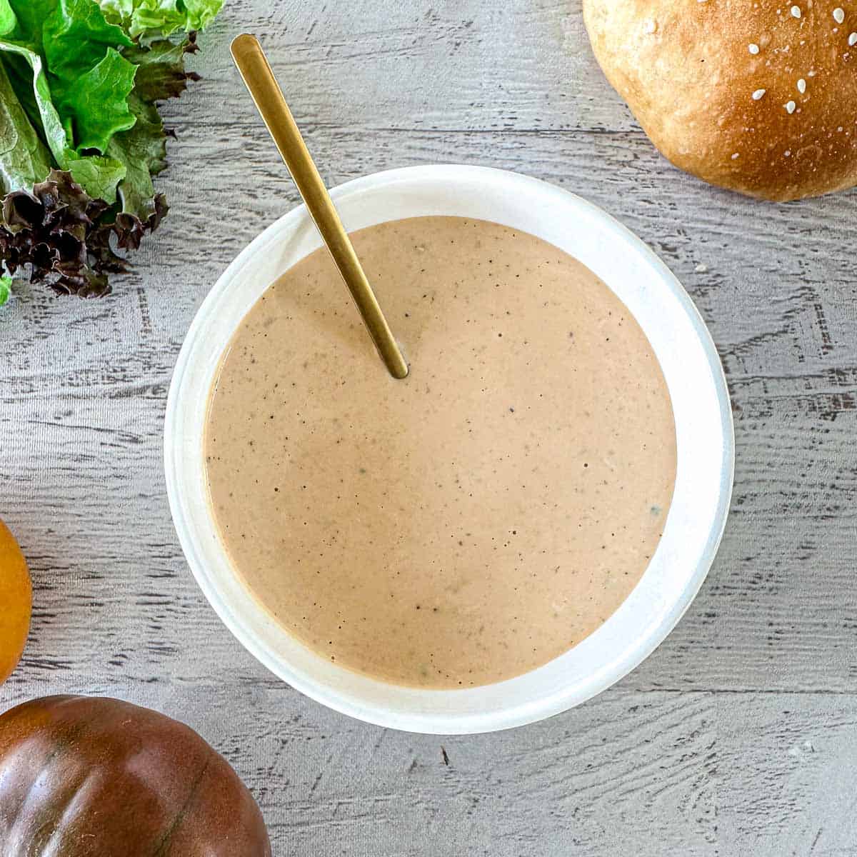A bowl of creamy, light brown sauce with a spoon inside sits on a wooden surface, surrounded by lettuce, a hamburger bun, and heirloom tomatoes.