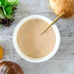A bowl of In-N-Out sauce with a spoon in it, placed on a wooden surface. Fresh lettuce, a sesame sandwich bun, and two tomatoes are arranged nearby.