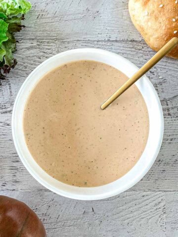 A bowl of In-N-Out sauce with a spoon in it, placed on a wooden surface. Fresh lettuce, a sesame sandwich bun, and two tomatoes are arranged nearby.
