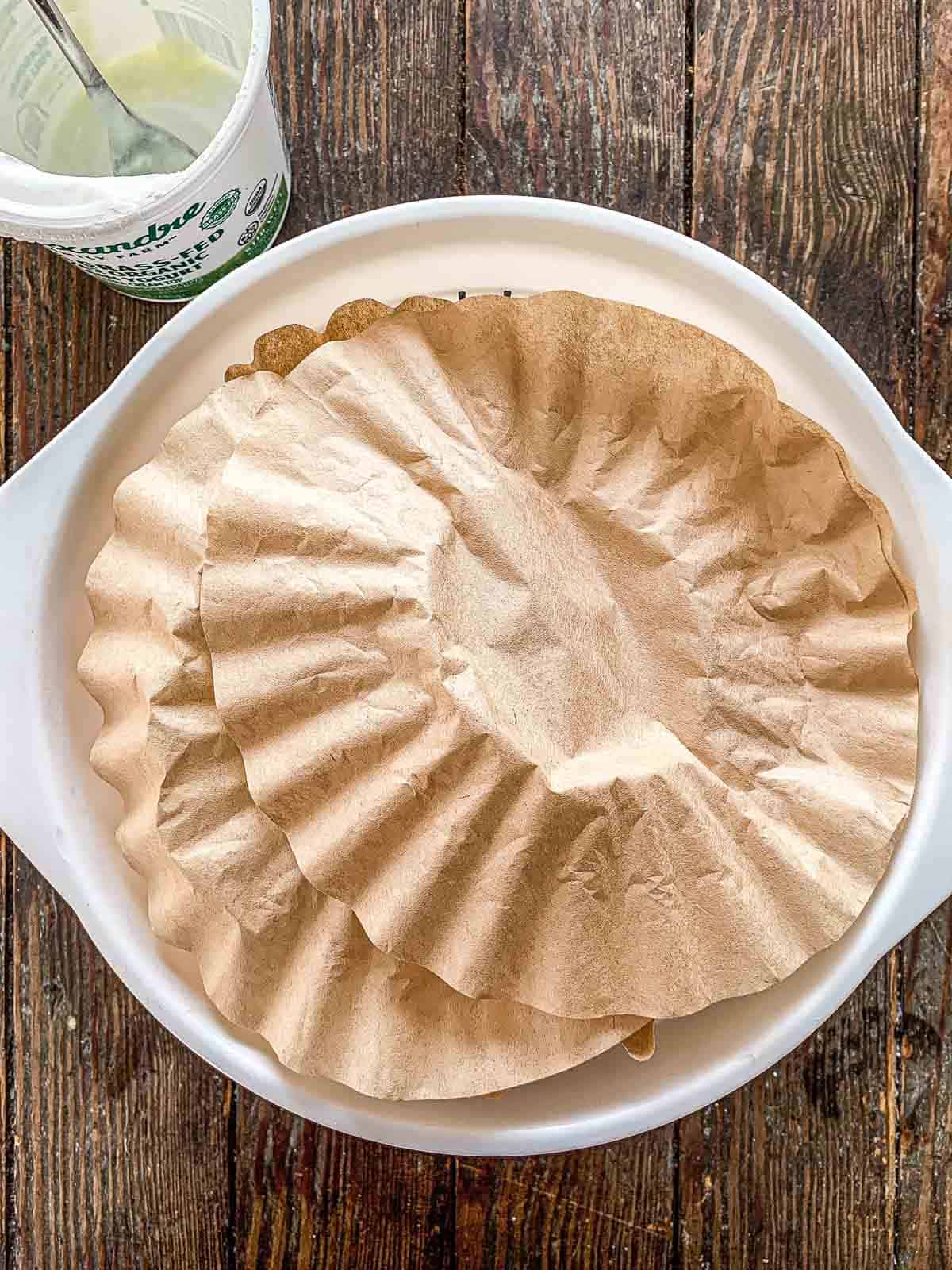 Two brown coffee filters are stacked in a white bowl, with a plastic container partially visible in the upper left corner. The bowl rests on a wooden surface.