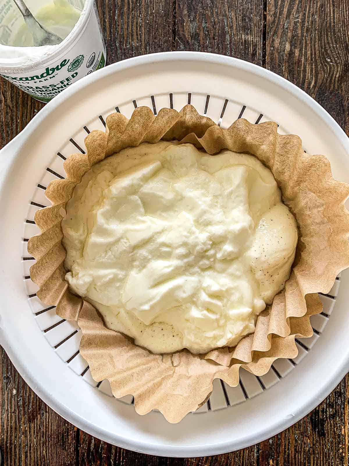 A round colander lined with brown parchment paper holds creamy yogurt cheese. A container of plain yogurt sits beside it on a rustic wooden surface.