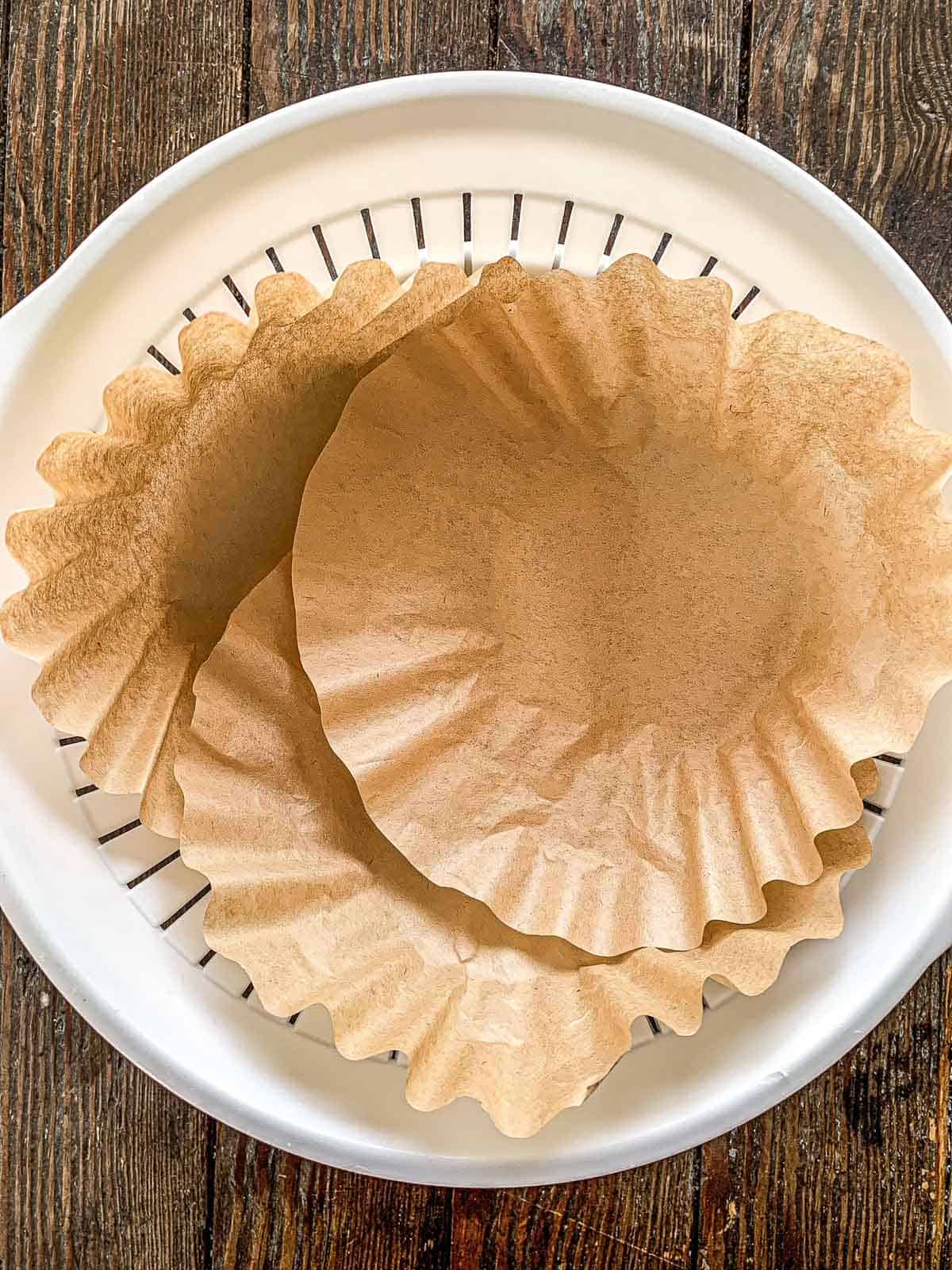 Two brown coffee filters placed inside a white plastic colander, all set on a wooden surface.