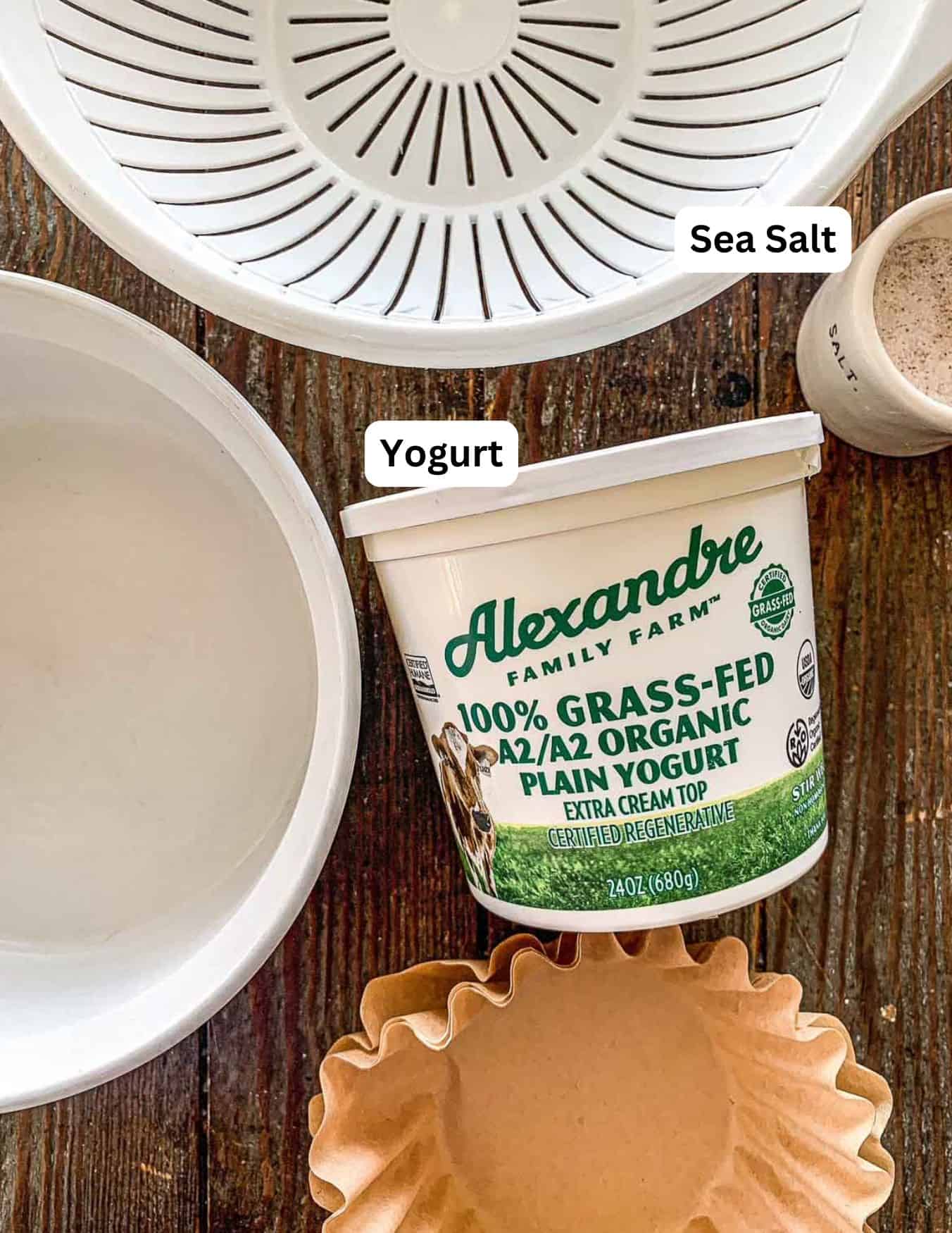 A container of organic plain yogurt, a bowl, a strainer, a small bowl labeled sea salt, and a brown coffee filter are arranged on a wooden surface.