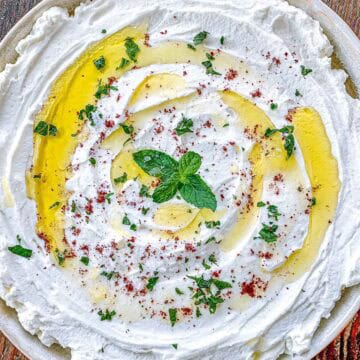 A plate of whipped labneh drizzled with olive oil, sprinkled with chopped herbs and spices, and garnished with a sprig of fresh mint.