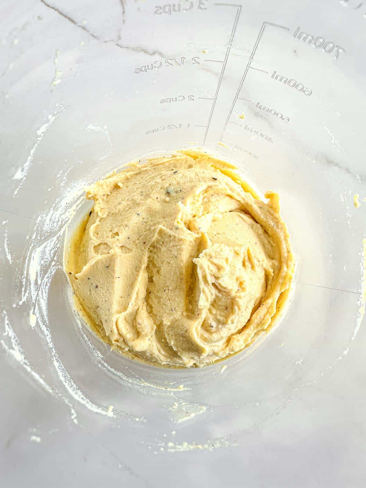 Thick, pale yellow batter is spread inside a clear mixing bowl with measurement marks on the side, sitting on a white countertop.