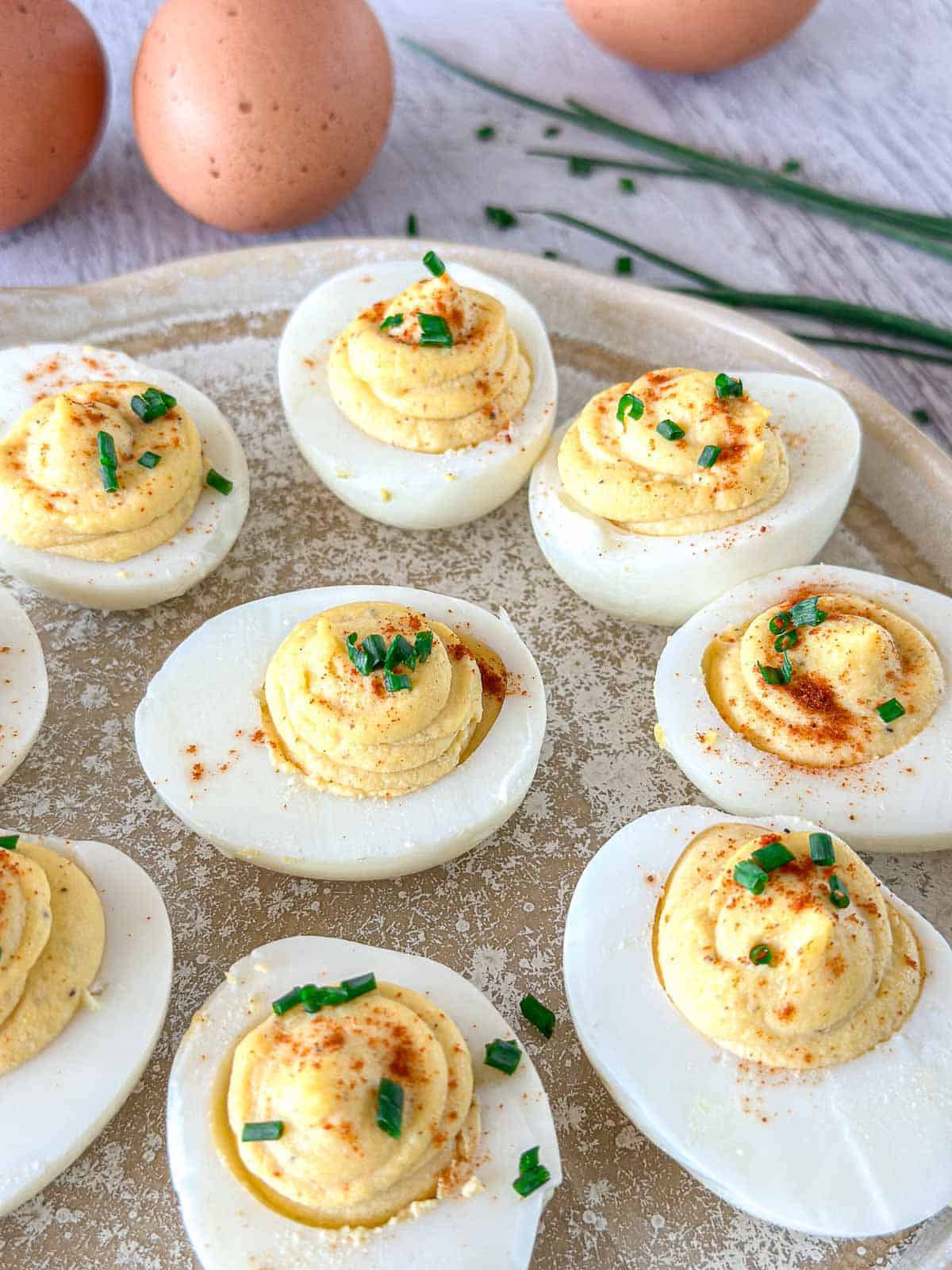 A plate of deviled eggs garnished with chopped chives and paprika, with whole eggs and fresh chives in the background.