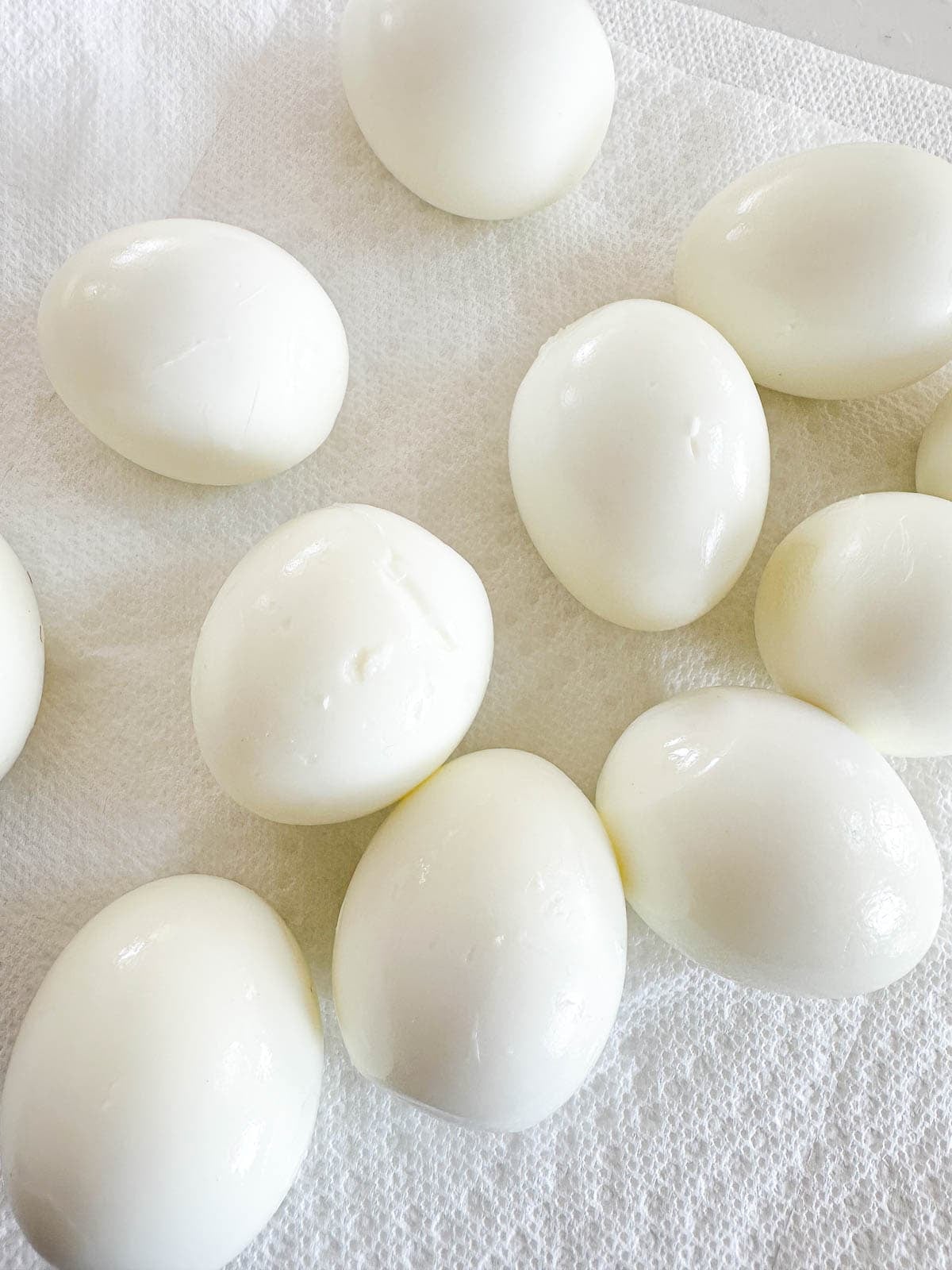 Several peeled hard-boiled eggs lie on a white paper towel, with smooth, shiny surfaces and a few small cracks visible on some of the eggs.
