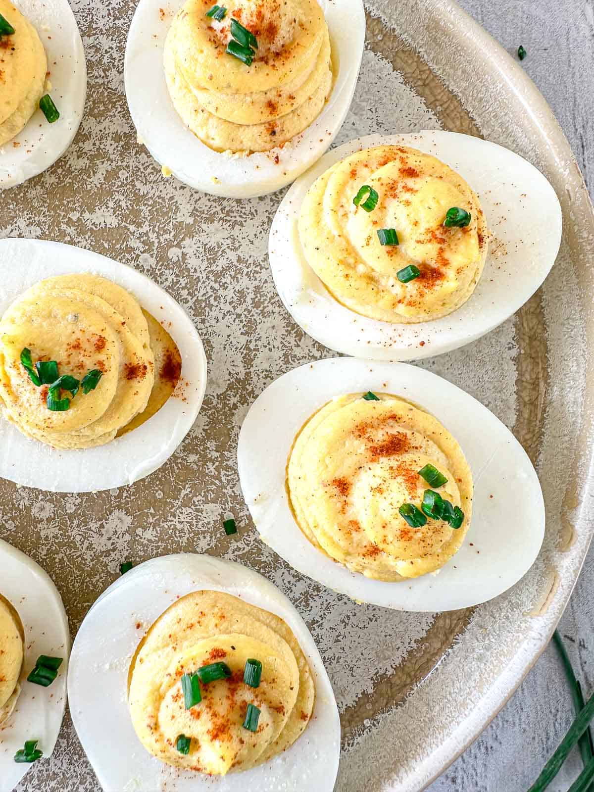 A platter of smoked deviled eggs topped with paprika and chopped chives sits on a textured plate. The eggs are halved, filled with creamy yolk mixture, and arranged neatly.