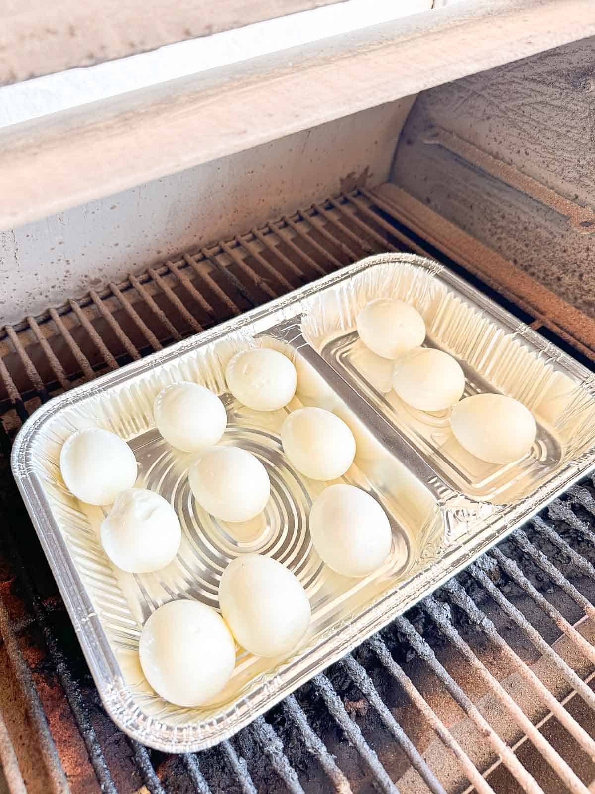 A disposable aluminum tray with ten peeled hard-boiled eggs sits on a grill grate, ready to be smoked.